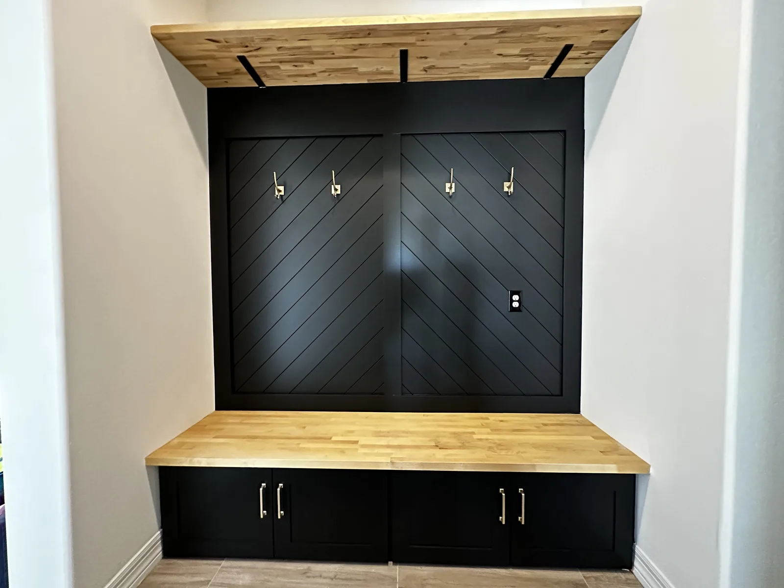 Modern black built-in entertainment center with diagonal shiplap backing, wooden top and base, four white hooks, electrical outlet, and cabinet storage below
