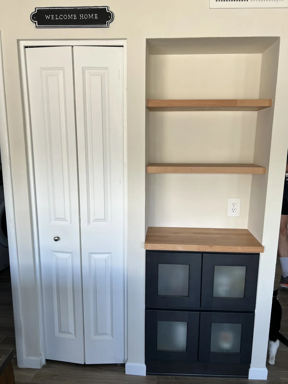 White closet doors next to built-in shelving unit with wooden shelves and black cabinet base with glass doors in home entryway with "Welcome Home" sign
