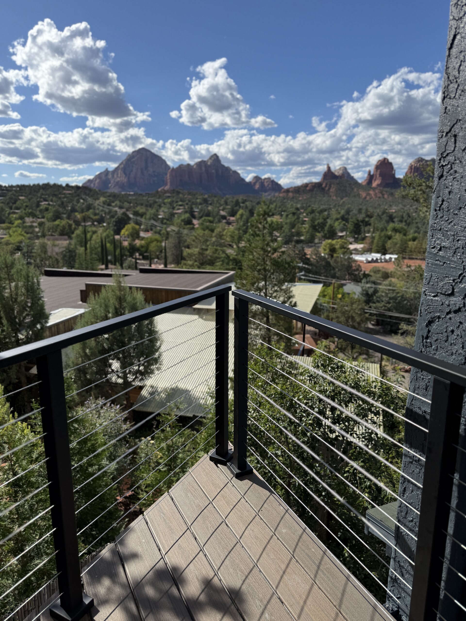 Modern deck with black cable railing overlooking red rock formations and forested landscape under blue sky with white clouds