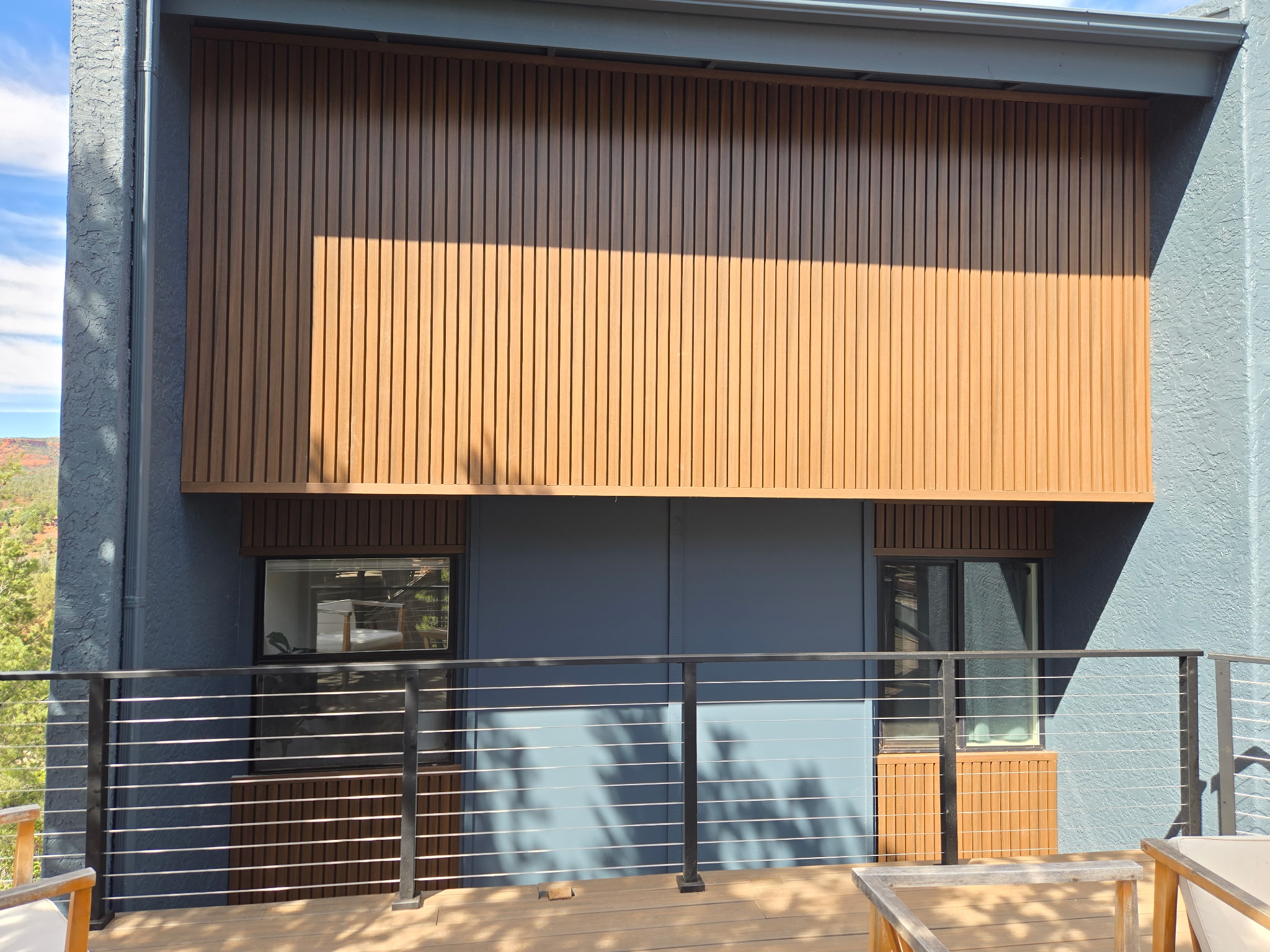 Modern residential building exterior with wood-slat sunshade, dark metal railings, and large windows overlooking green landscape