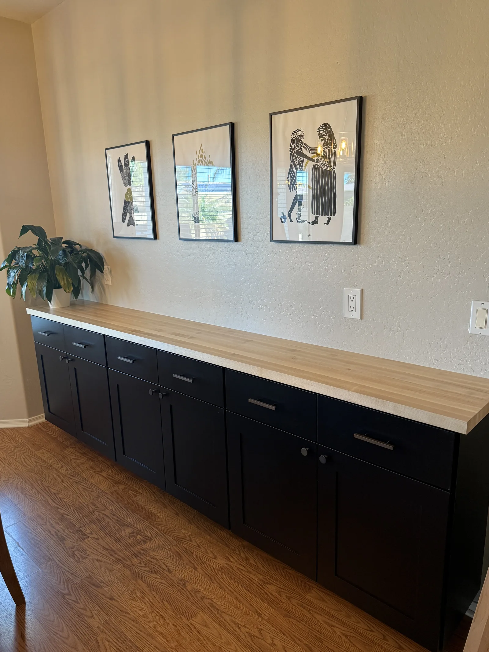 Black cabinetry with light wood countertop displaying potted plant, three framed line drawings on cream wall above, hardwood flooring visible below