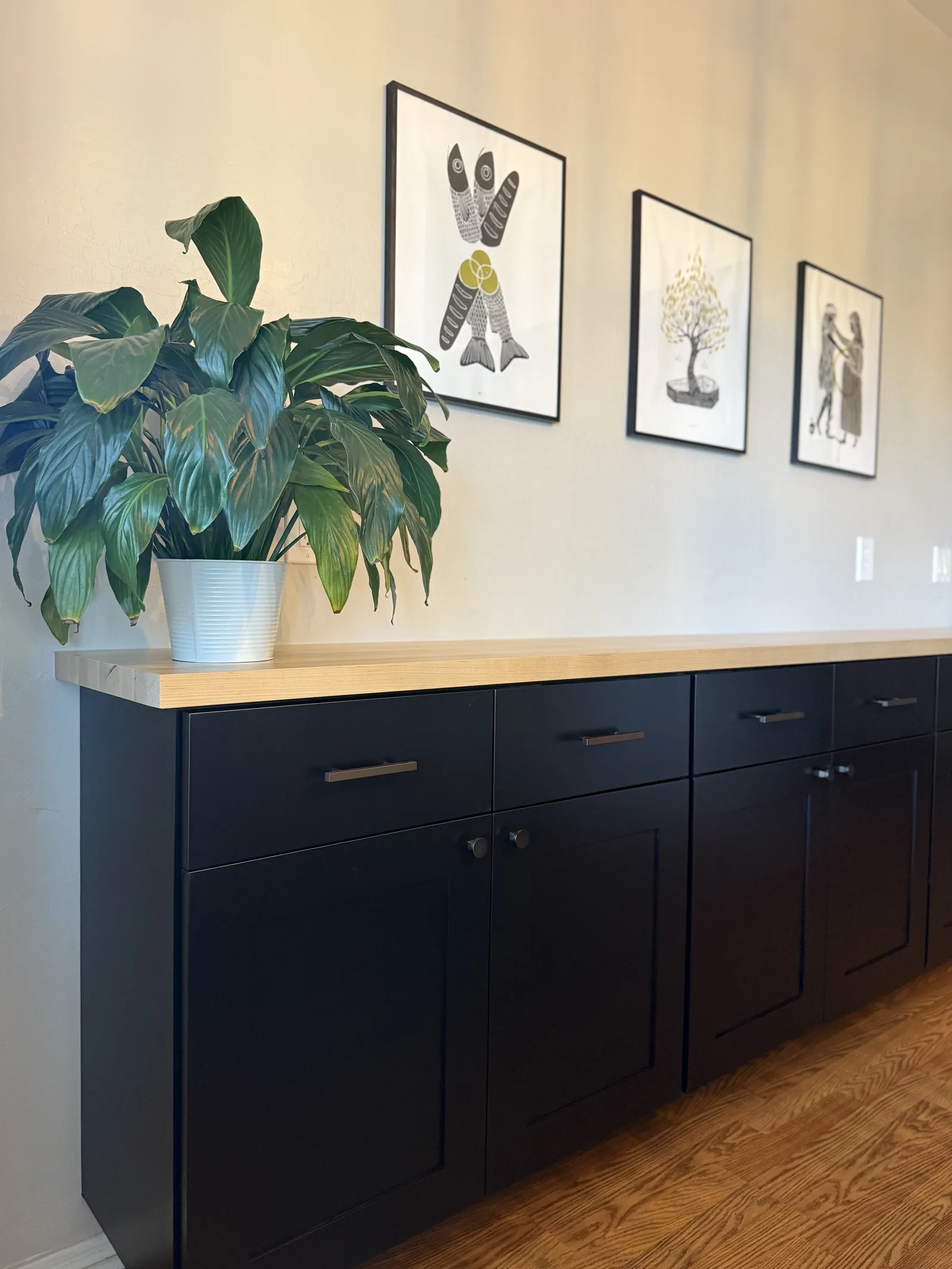 Black cabinet with light wood countertop displaying potted plant and three framed botanical illustrations on cream wall
