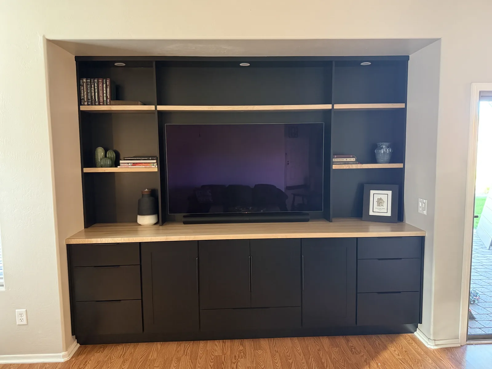 Modern built-in entertainment center with dark cabinetry, light wood shelving, and flat-screen TV mounted in alcove, featuring decorative items and books on open shelves above