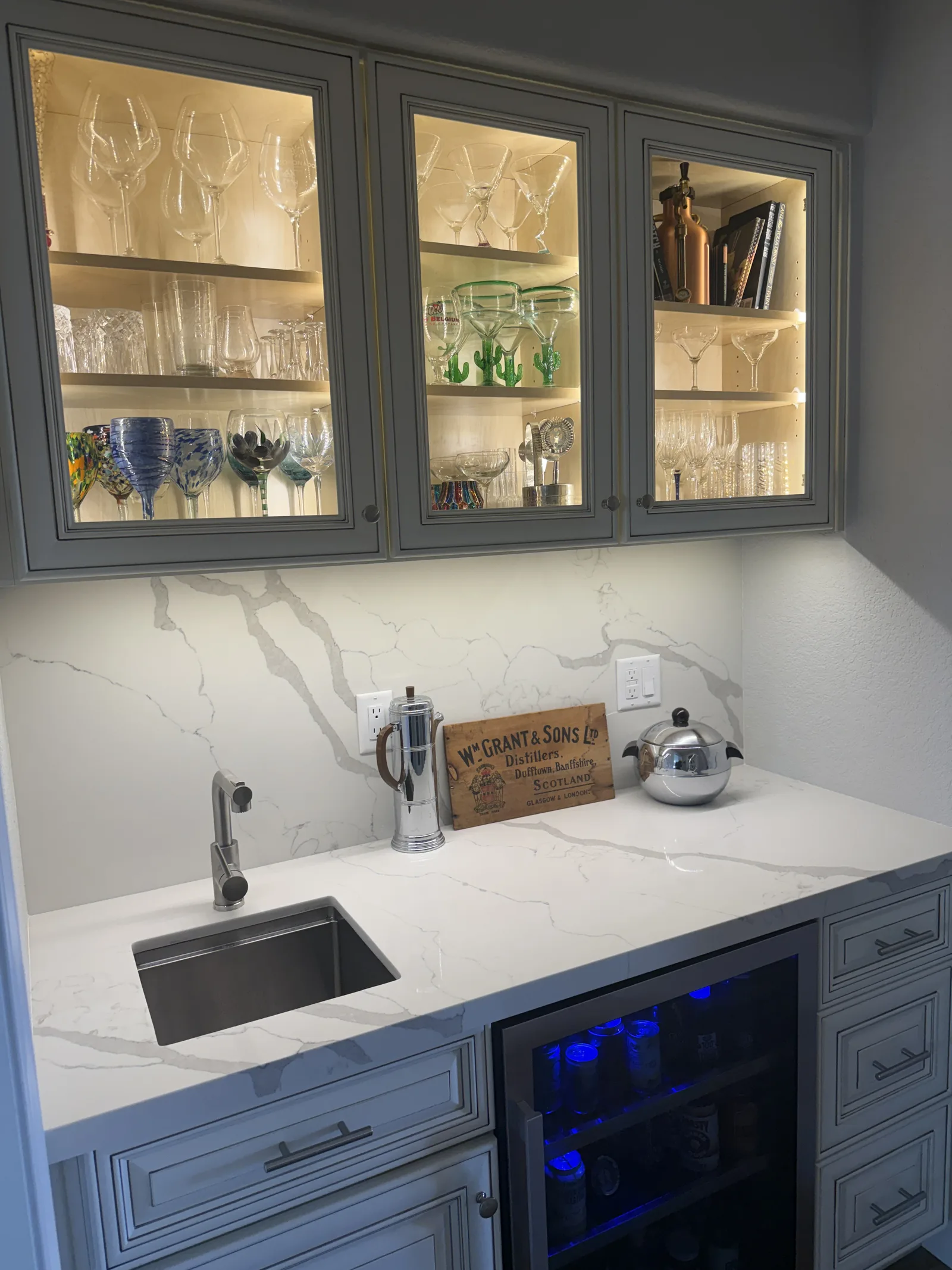 Elegant wet bar with gray cabinetry, marble countertop, glass-front upper cabinets displaying wine glasses and spirits, blue-lit wine cooler, and decorative whisky box