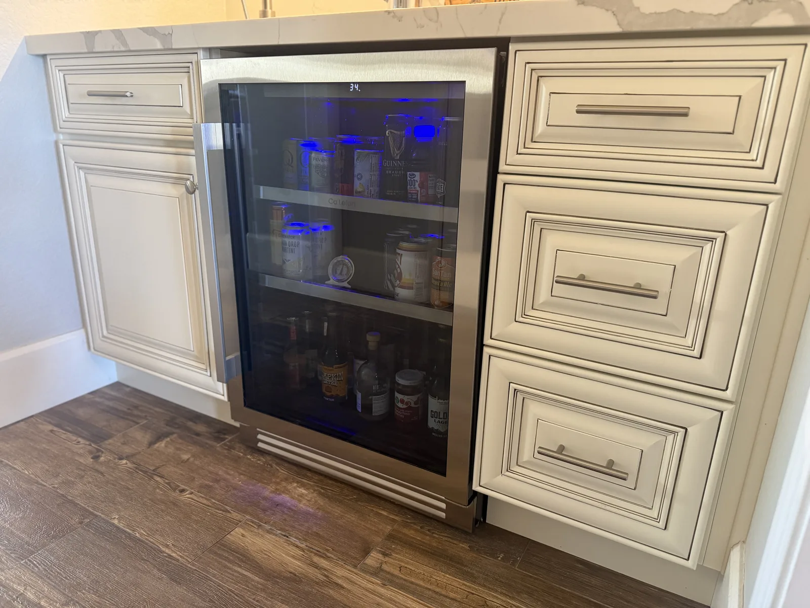 Built-in beverage cooler with glass door and blue LED lighting, flanked by cream-colored cabinetry with drawers, installed in kitchen with wood flooring