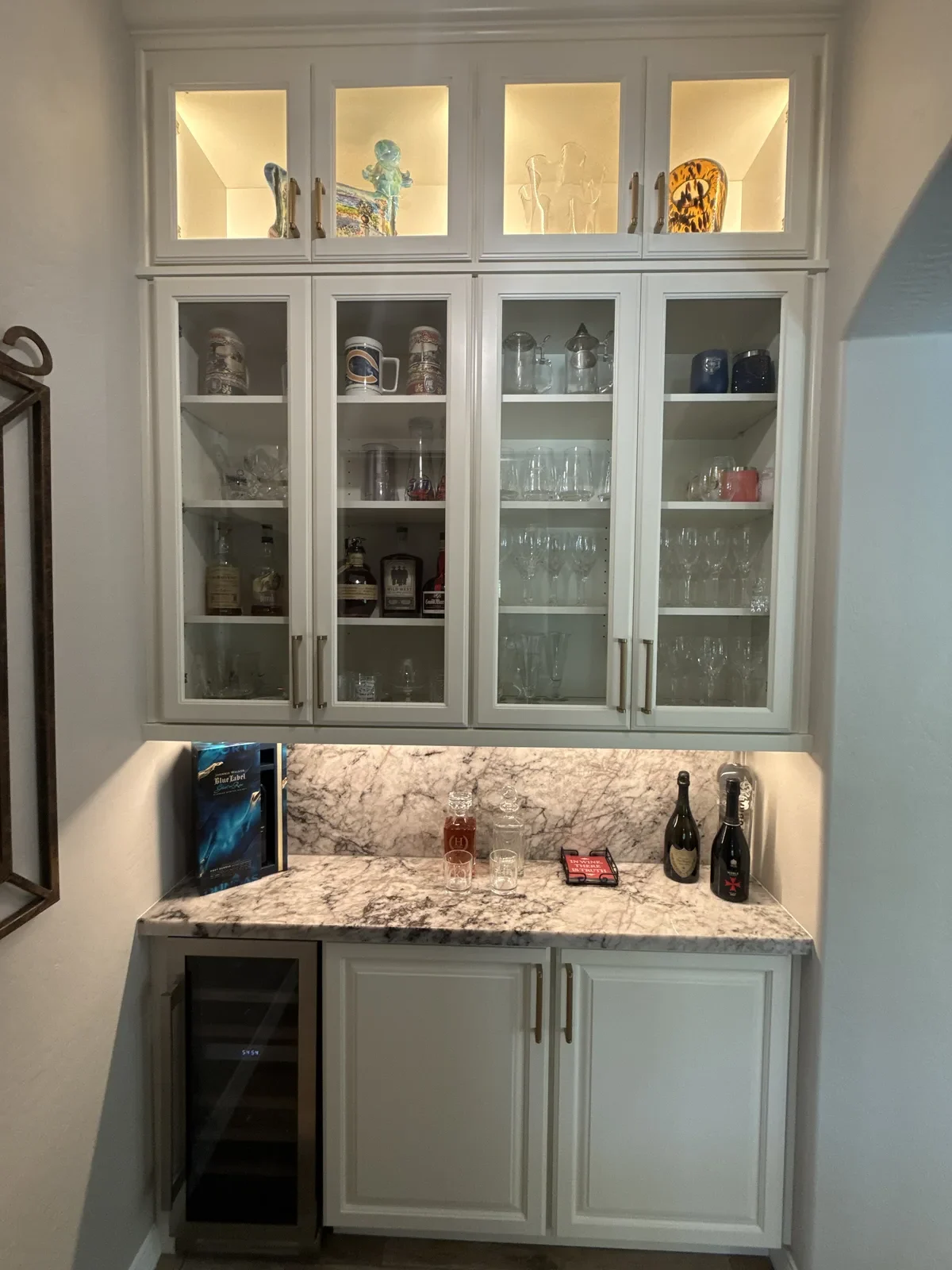 Built-in bar cabinet with white frame, glass-front upper shelves displaying decorative items and glassware, marble countertop with wine bottles and bar accessories, and wine cooler on lower left
