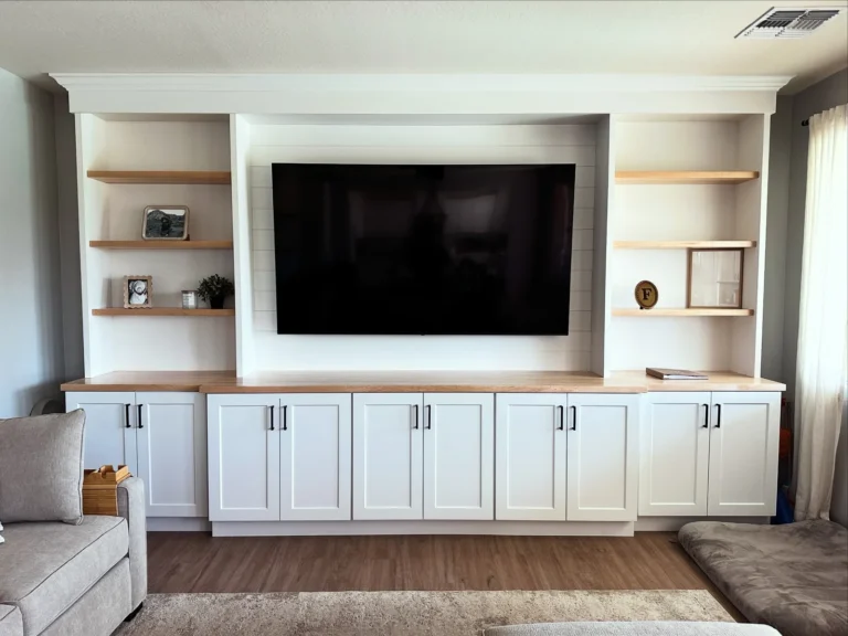Modern living room with built-in white cabinetry, wooden shelving, and mounted flat-screen TV between two accent chairs on hardwood flooring