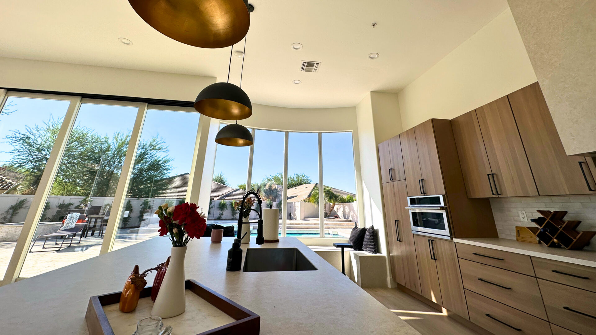 Modern kitchen with white countertops, wood cabinetry, and black pendant lights overlooking a backyard pool through floor-to-ceiling windows