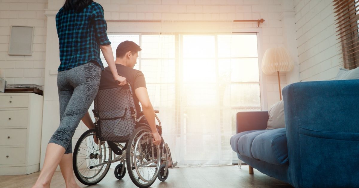 Two people in a bright, modern living room with large windows; one person stands behind a wheelchair-using individual, both looking toward the sunlit doorway ahead