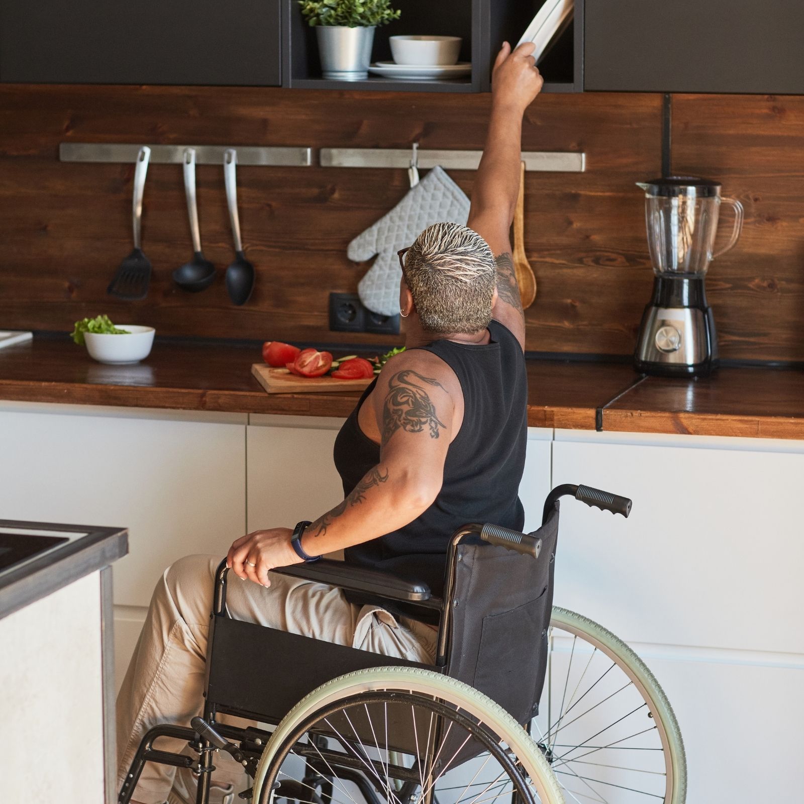 Woman in wheelchair reaching for items in accessible kitchen with wooden countertop, preparing food with fresh vegetables and blender nearby