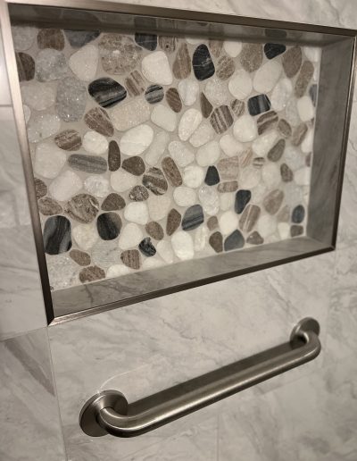 Accessible bathroom shower with stainless steel ADA grab bar, built-in recessed niche, pebble stone mosaic tile, and marble-look wall tile
