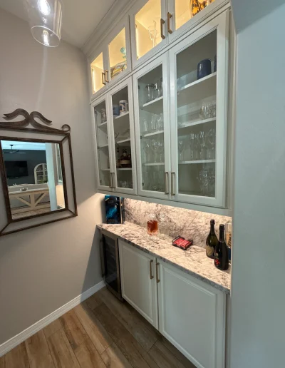 Custom dry bar with white glass-front cabinetry, illuminated display cabinets, stone countertop, beverage refrigerator, and brass hardware