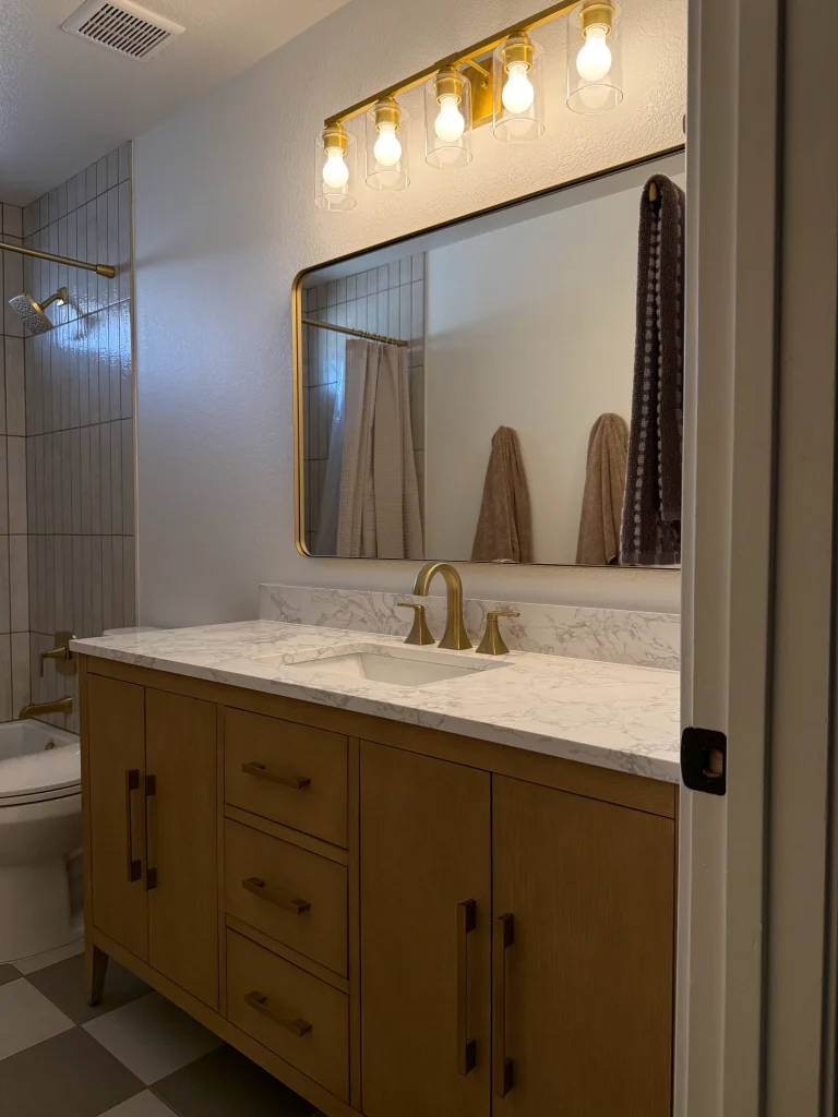Remodeled guest bathroom with wood vanity, marble-look countertop, brushed gold fixtures, large framed mirror, and vertical shower tile
