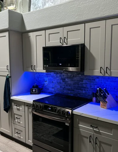 Kitchen remodel with gray shaker cabinets, white countertops, stainless steel appliances, dark stone backsplash, and blue under-cabinet lighting