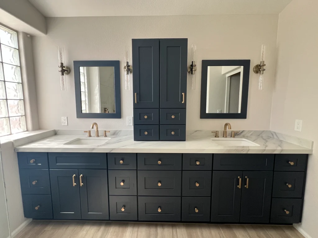 Primary bathroom remodel with dark blue double vanity, marble-look countertop, brass fixtures, framed mirrors, and matching built-in storage
