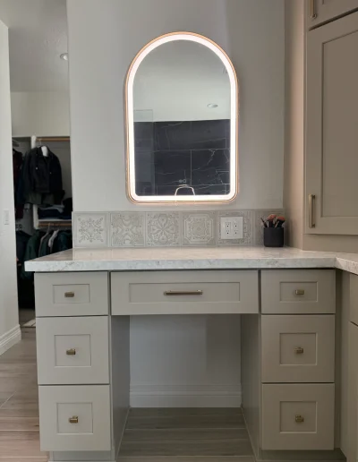 Custom bathroom makeup vanity with taupe cabinetry, quartz countertop, decorative tile backsplash, and illuminated arched mirror