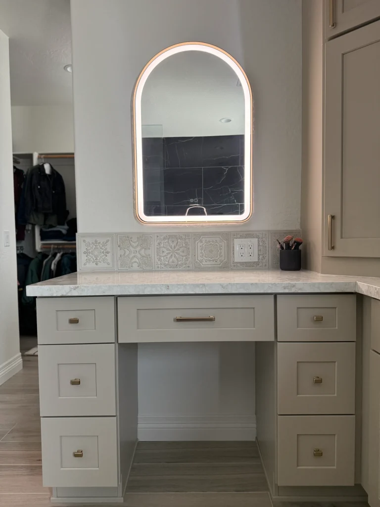 Custom bathroom makeup vanity with taupe cabinetry, quartz countertop, decorative tile backsplash, and illuminated arched mirror