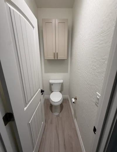 Remodeled private toilet room with custom wall-mounted storage cabinet, wood-look tile flooring, and white paneled door