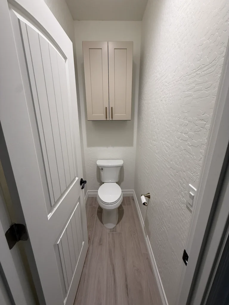 Remodeled private toilet room with custom wall-mounted storage cabinet, wood-look tile flooring, and white paneled door