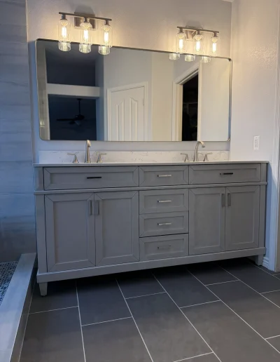 Remodeled primary bathroom with gray double vanity, quartz countertop, brushed nickel fixtures, large wall mirror, and modern vanity lighting