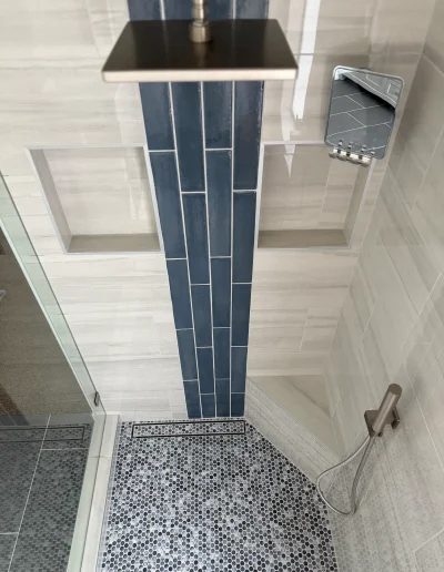 Custom walk-in shower with gray penny tile floor, white wall tile, blue accent tile, built-in niches, linear drain, and handheld shower fixture