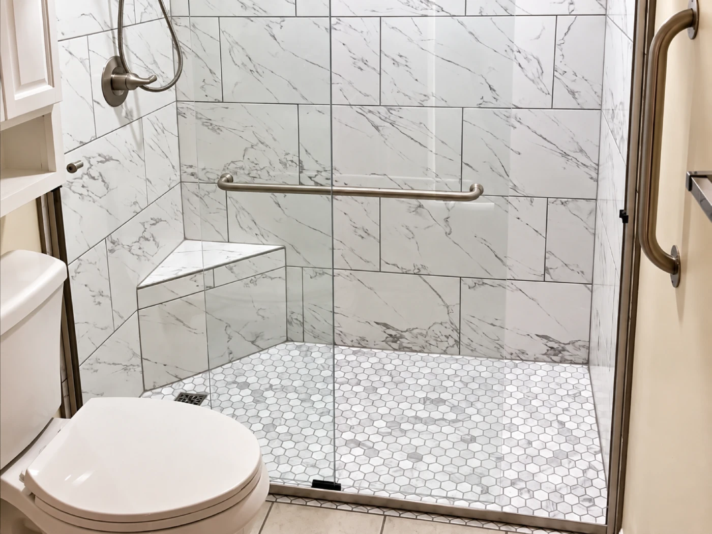 adu bathroom (1) Accessible walk-in shower remodel featuring marble-look wall tiles, hexagonal mosaic flooring, built-in shower bench, glass enclosure, and grab bars for added safety